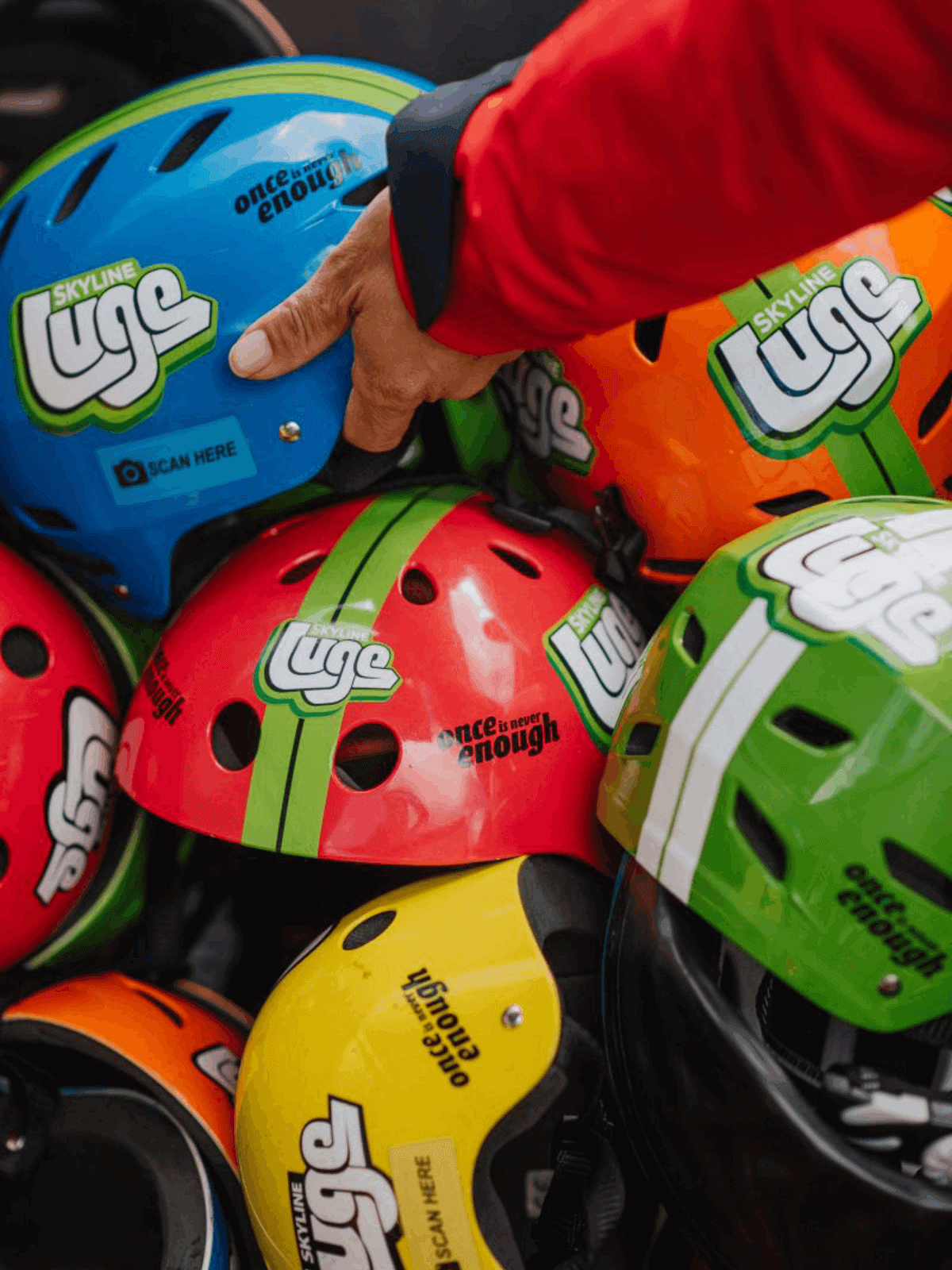 Close-up of colourful Skyline Luge helmets stacked together, with a staff member picking up a blue helmet.