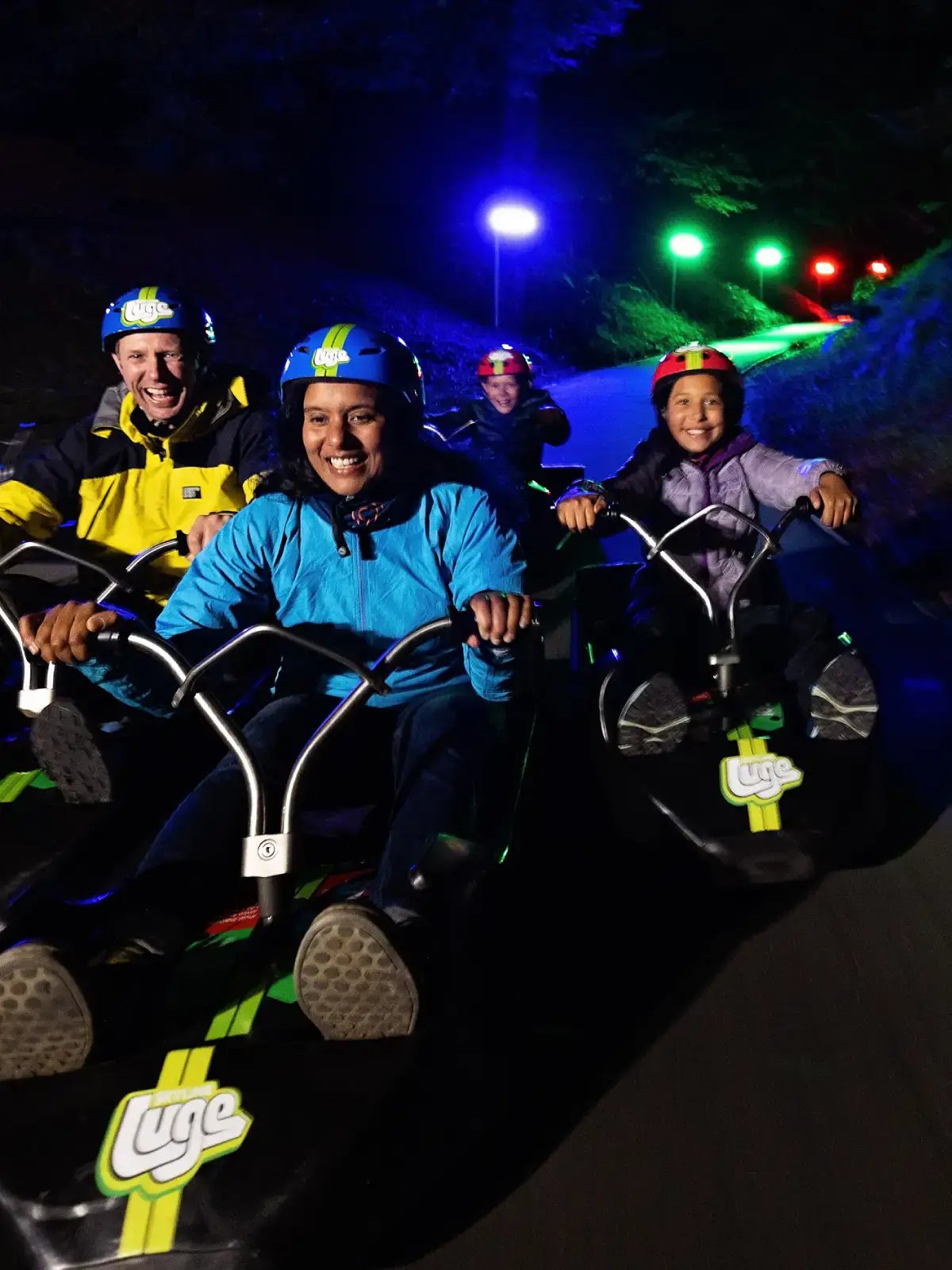 Family riding the Twilight Luge at Skyline Rotorua, smiling as they zoom under colourful lights at dusk.