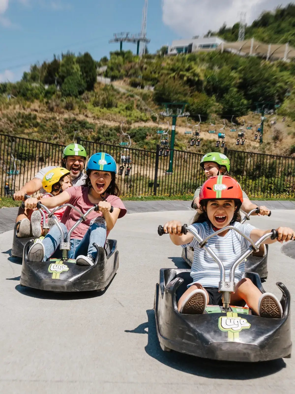 Kids and parents laughing as they Luge down the track at Skyline Rotorua on a sunny day.