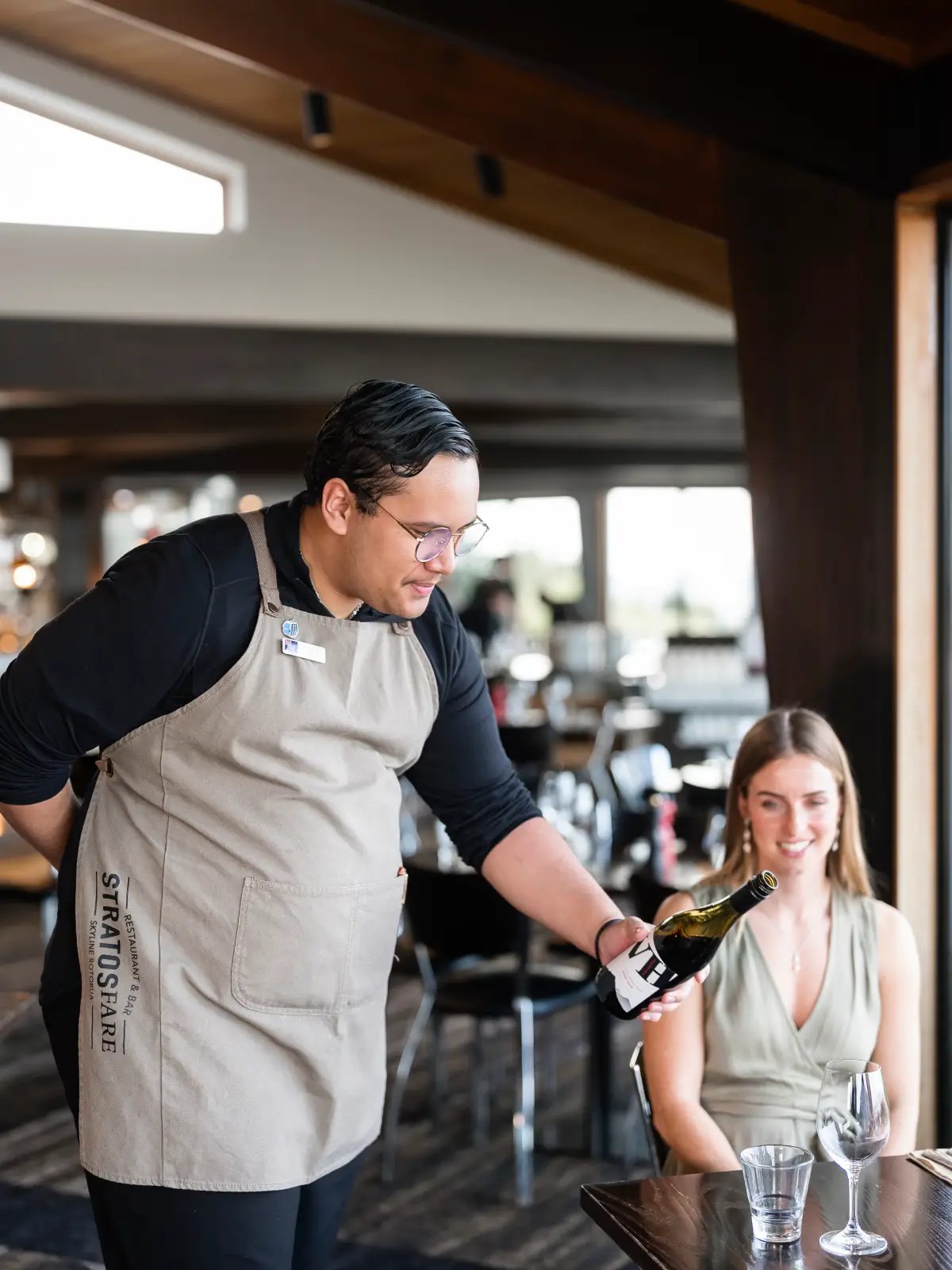 A Stratosfare staff member presents a bottle of wine to a seated guest inside the restaurant, with natural light shining through the window and tables set behind them.