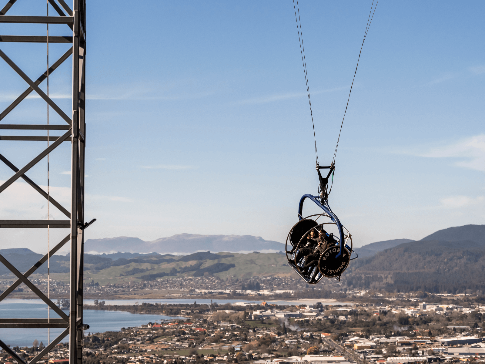 Three people on the Skyswing at Skyline Rotorua, soaring high above the city with lake views and Mount Tarawera in the background.