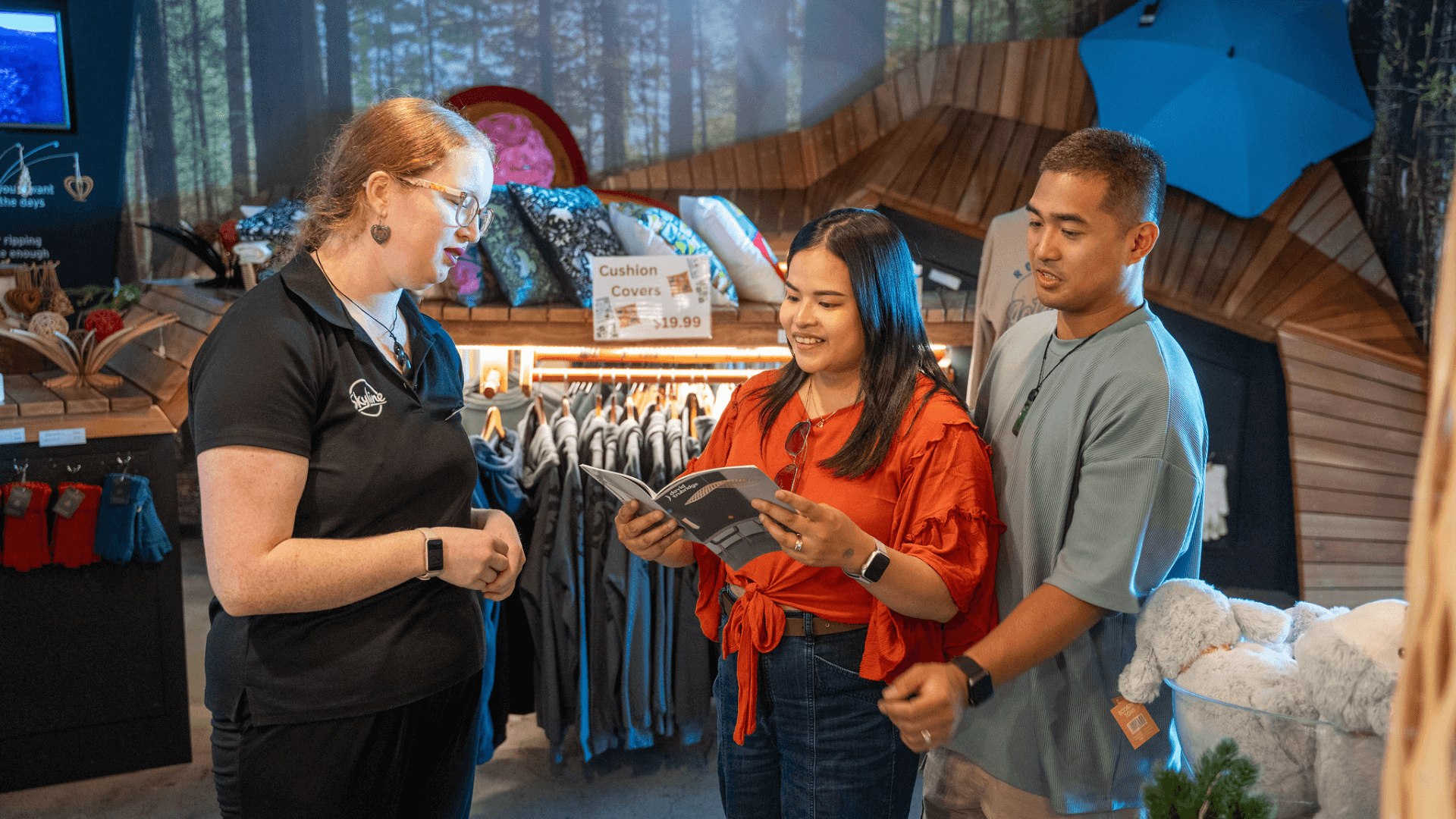 A Skyline Rotorua staff member assists two guests browsing products and gifts at the iNZpired store, smiling and chatting together.