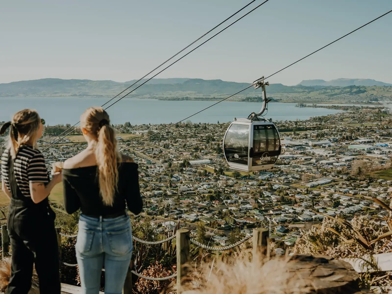 Two friends enjoying the view as a Skyline Rotorua gondola passes overhead with Lake Rotorua and the city below.