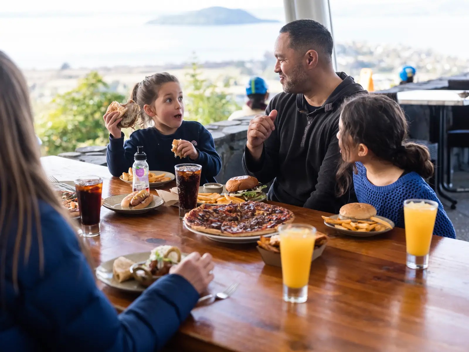 Family enjoying burgers, pizza and drinks at Market Kitchen Café at Skyline Rotorua with lake views in the background.