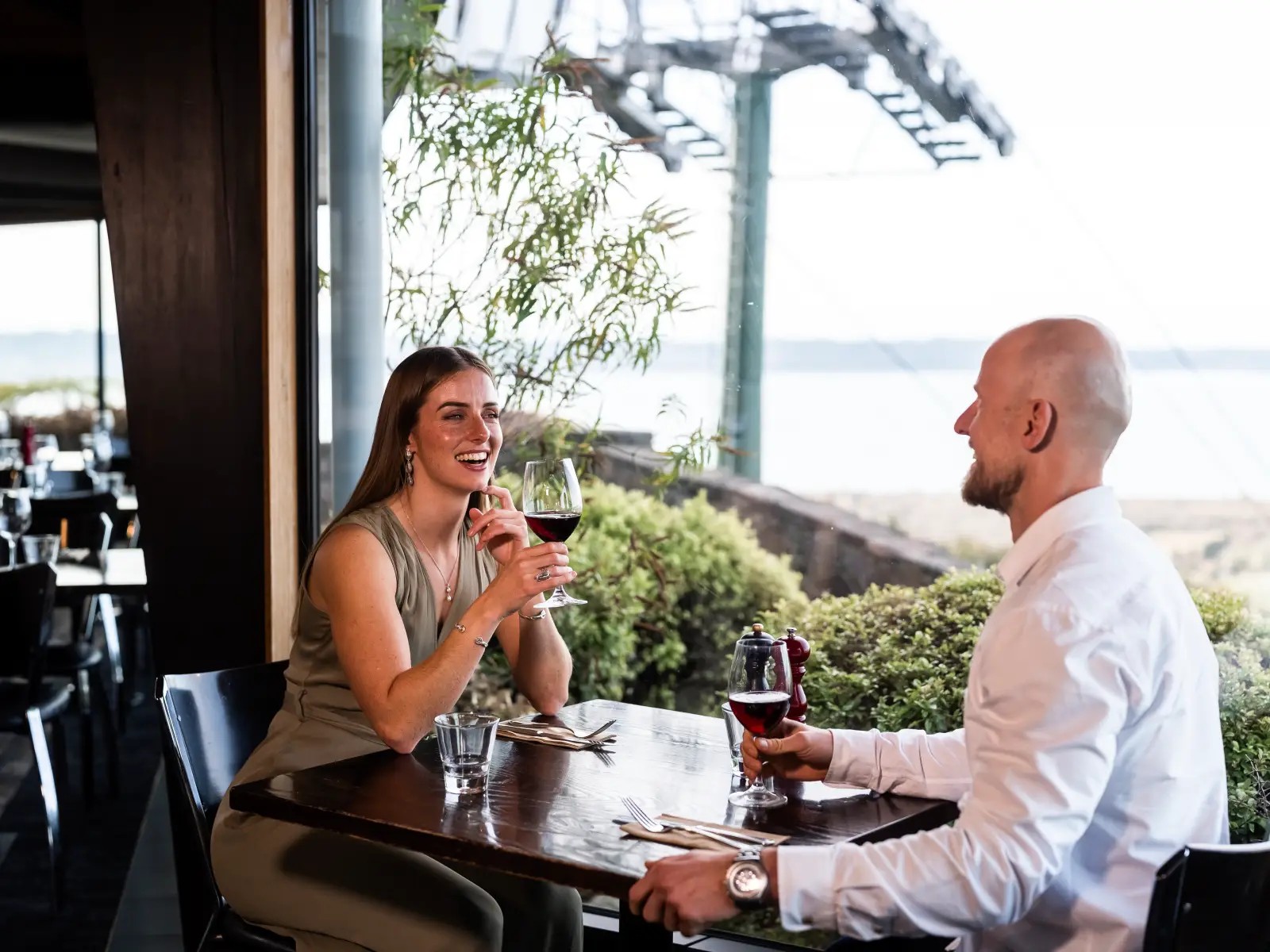 A couple enjoys glasses of red wine at a window table in Stratosfare, with views overlooking Rotorua and the Gondola structure outside.