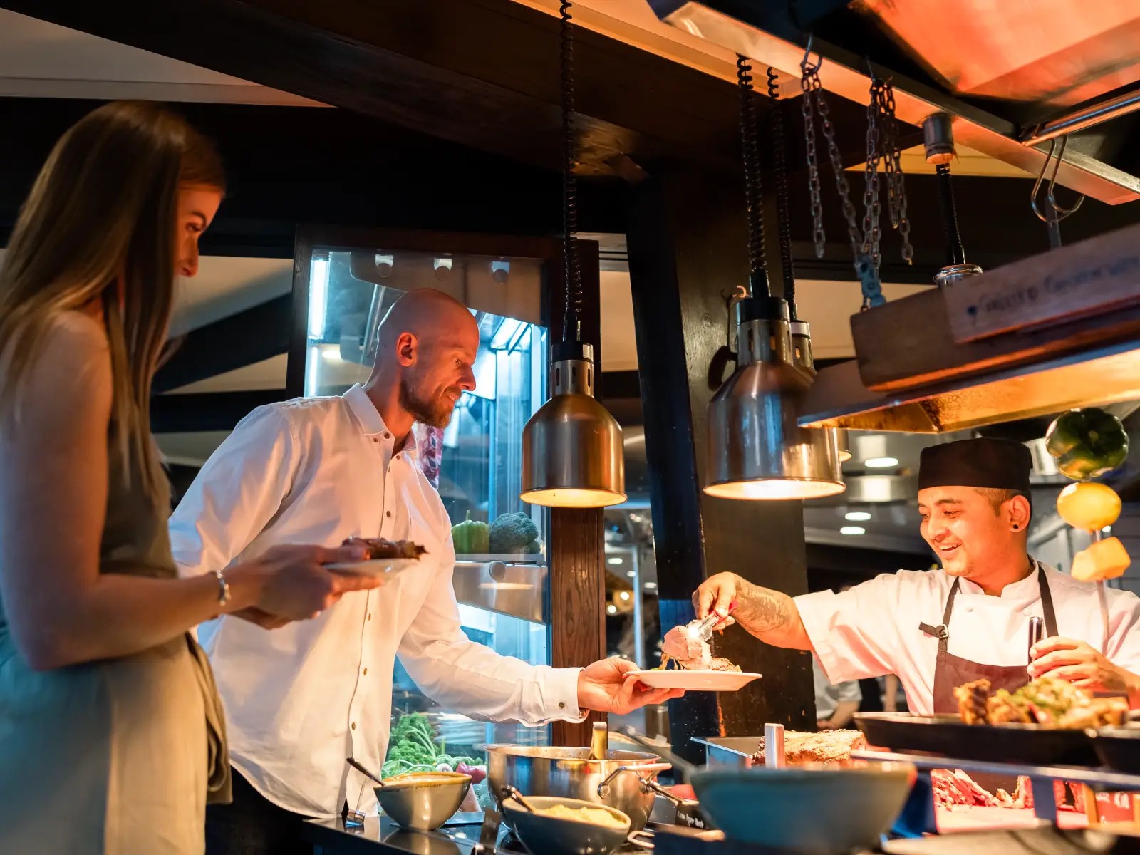 A Stratosfare chef serves food to a couple at the buffet station in the evening, with warm lighting and views of the restaurant behind them.
