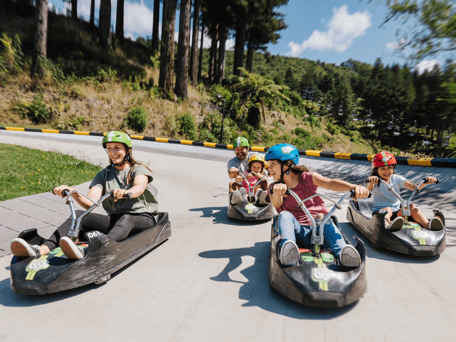 Family zooming down the Luge track at Skyline Rotorua on a sunny day, smiling and enjoying the ride through the forest.