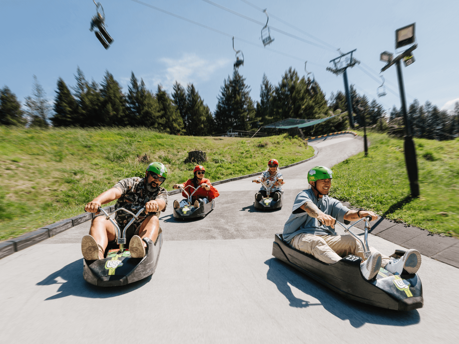Group of adults zooming down the Luge track at Skyline Rotorua under the summer sun, surrounded by forest and chairlift views.